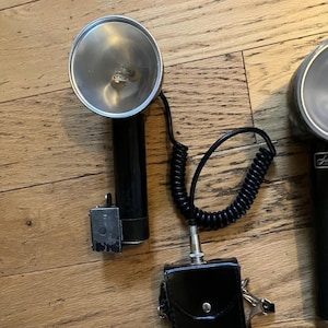 May include: Vintage Sun-Lite flash unit with a black handle and a silver reflector. The unit is connected to a black battery pack with the text "SUN-LITE Battery Pac". A coiled black cord connects the flash to the battery pack.