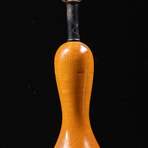 May include: A table lamp featuring a polished light brown wooden base shaped like a bowling pin, topped with a brass socket and a white frosted glass shade. The lamp stands on a circular black base with a black power cord wrapped around the bottom.