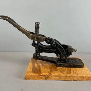 May include: An antique metal button maker with a bronze lever and a black base, mounted on a wooden block. The lever is curved and connected to a spring. The base has text.