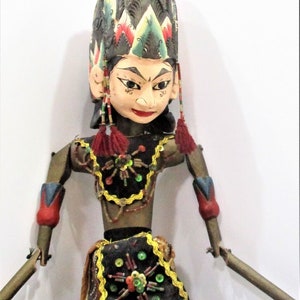May include: A wooden puppet with a colourful headdress, a black and gold beaded bodice, and a brown and gold skirt with colourful beads. The puppet has a painted face with red lips and black eyes.