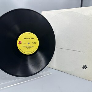 May include: A black vinyl record with a yellow label featuring text and a red tongue logo. The record is next to a white album cover with text and a Rolling Stones logo. The record is displayed on a clear stand.