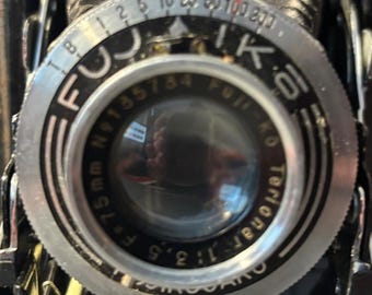 Peut inclure: Gros plan sur l'objectif d'un appareil photo Fuji vintage. L'objectif a une lunette argentée avec des marquages noirs pour l'ouverture et la mise au point. L'objectif est marqué "75mm f:3.5" et "Fujinar 1:3.5". Le corps de l'appareil photo est noir.