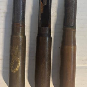 May include: Three cylindrical metal artillery shells arranged vertically. The left shell features a faded gold-colored emblem. The center shell has a hollowed-out section revealing internal threading. All three shells have dark, weathered casings.