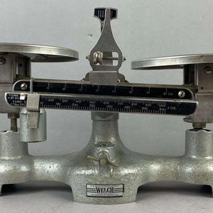Könnte beinhalten: Eine vintage Welch Balkenwaage mit einem silberfarbenen Metallsockel und zwei runden Waagschalen. Der zentrale Balken verfügt über schwarze Skalen mit weißen Markierungen, einschließlich Angaben in Unzen und Gramm, sowie einen vertikalen Zeiger.