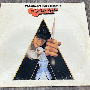 May include: A vintage vinyl record album cover featuring a black and white image of a man holding a knife in a triangular frame. The album title "Clockwork Orange" is printed in white with a red and orange circle behind it. The album is by Stanley Kubrick.