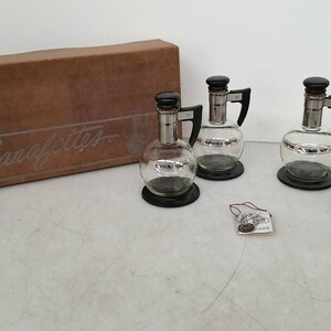 May include: Set of four glass carafe decanters with black lids and handles. The decanters have a silver band around the middle and are sitting on black bases. The decanters are in a brown box with the word "Carafelettes" printed on it.