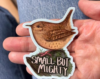 May include: A sticker featuring a detailed illustration of a small brown bird perched on a mossy rock. The text "SMALL BUT MIGHTY" is written in a stylized font below the bird. The sticker has a light blue border.