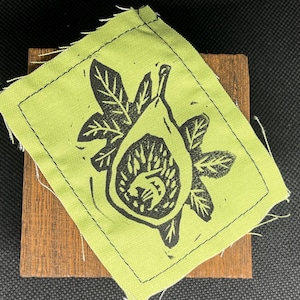 Puede incluir: Un parche de tela verde lima con un grabado en linóleo negro de un higo y hojas. El parche está cosido con hilo negro y descansa sobre un pequeño bloque de madera rectangular. El fondo es una superficie oscura y texturizada.