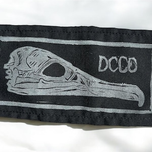 May include: Black rectangular patch featuring a detailed silver illustration of a bird skull. The skull is centered within a silver border, with the letters "DCC0" in the upper right corner. The patch is made of a dark fabric.