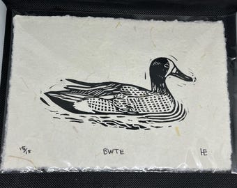 May include: A black and white linocut print of a duck on water. The artwork is on textured paper and includes the text "BWTE" and "15/5". The print is in a protective sleeve.