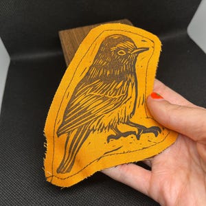 May include: An orange fabric patch featuring a detailed black linocut print of a bird. The bird is depicted in profile, with intricate feather details and a watchful eye. The patch has a stitched border and is held in a hand.