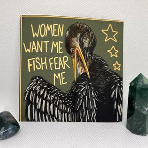 Peut inclure: Une carte verte avec le texte "Women want me Fish fear me" en lettres dorées. Un oiseau noir avec un long bec jaune est illustré sur la carte. Il y a trois étoiles dorées dans le coin supérieur droit de la carte.