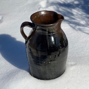 May include: A dark brown ceramic pitcher with a handle and a spout. The pitcher has a glossy finish and a slightly textured surface. The interior is a lighter brown. The pitcher is sitting on a bed of snow.