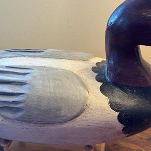May include: A hand-carved wooden duck decoy. The duck has a white body with gray and black painted details. The head and neck are a deep burgundy color. The decoy is a decorative item.