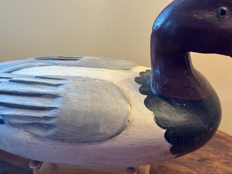 May include: A hand-carved wooden duck decoy. The duck has a white body with gray and black painted details. The head and neck are a deep burgundy color. The decoy is a decorative item.