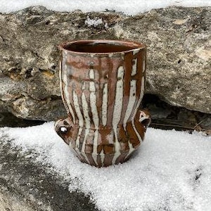 May include: A handmade ceramic vase with a brown and white glaze. The vase has a cylindrical shape with two small handles and a textured surface. The glaze drips down the sides, creating a unique pattern. The vase is approximately 6 inches tall.
