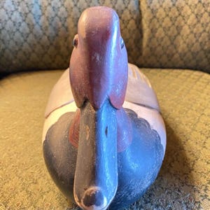 May include: A hand-carved wooden duck figurine. The duck has a purple head and beak, with a red patch on its neck. The body is painted in shades of gray, blue, and white. The duck is a decorative item, likely for display.