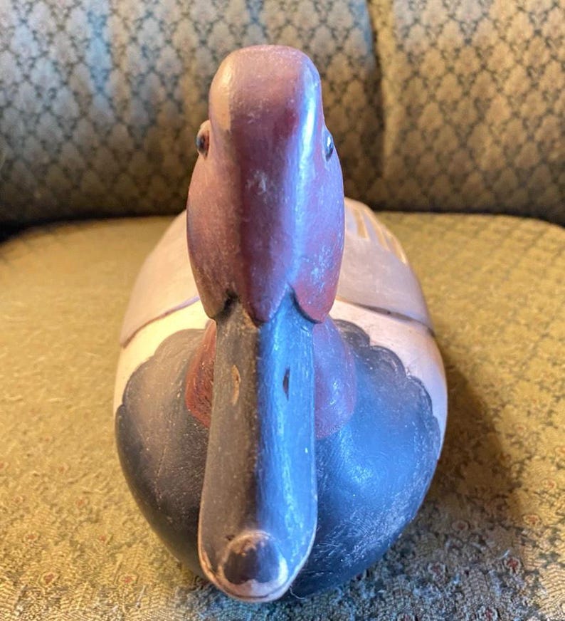 May include: A hand-carved wooden duck figurine. The duck has a purple head and beak, with a red patch on its neck. The body is painted in shades of gray, blue, and white. The duck is a decorative item, likely for display.