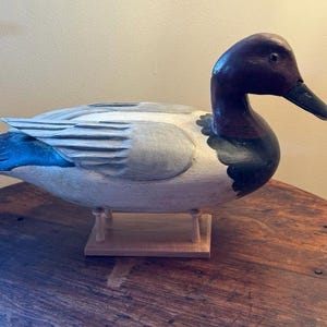 May include: A hand-carved wooden duck decoy with a detailed design. The duck features a dark burgundy head and neck, a black beak, and a light gray body with blue accents on the wings. It is mounted on a wooden base.