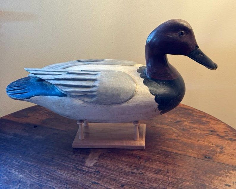 May include: A hand-carved wooden duck decoy with a detailed design. The duck features a dark burgundy head and neck, a black beak, and a light gray body with blue accents on the wings. It is mounted on a wooden base.