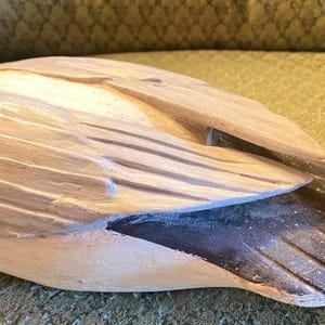 May include: A carved wooden duck figurine with a detailed wing design. The duck has a light brown body, a black tail, and a red head. The figurine is resting on a textured surface.