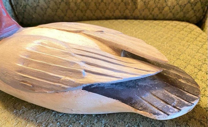 May include: A carved wooden duck figurine with a detailed wing design. The duck has a light brown body, a black tail, and a red head. The figurine is resting on a textured surface.