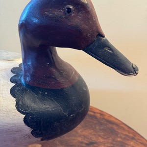 May include: A wooden duck decoy with a detailed, hand-painted design. The duck features a dark burgundy head and neck, a black beak, and a white body with black accents. The decoy is likely used for hunting or as a decorative item.