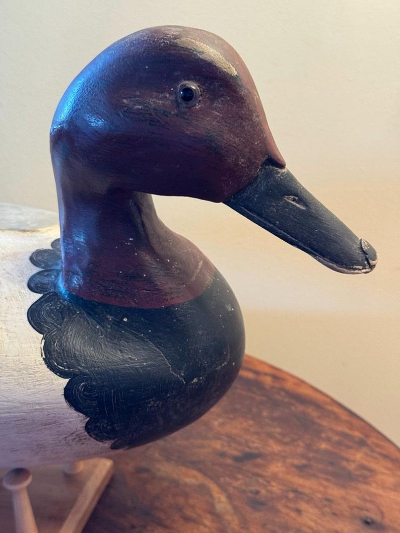 May include: A wooden duck decoy with a detailed, hand-painted design. The duck features a dark burgundy head and neck, a black beak, and a white body with black accents. The decoy is likely used for hunting or as a decorative item.