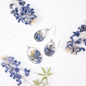 May include: A silver necklace with an oval pendant and a pair of matching teardrop earrings, each containing pressed blue and white lupine flowers encased in clear resin. The jewelry set is arranged alongside several loose sprigs of blue lupine flowers.