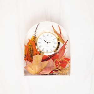 May include: An arched clear resin desk clock featuring embedded dried autumn leaves in shades of orange, red, and yellow. The clock face is circular with a gold-tone rim, white background, black Roman numerals, and black hour, minute, and second hands.