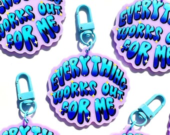 May include: A set of six purple keychains with a blue and white gradient design. Each keychain features the text "Everything Works Out For Me" in a bubbly font with glitter accents.