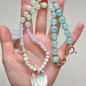 Peut inclure: Un collier de perles composé d'un mélange de perles opalescentes irisées, de perles vert pâle et de perles turquoise vif, rehaussé de petites entretoises dorées. Le collier comprend un rang de perles blanches et un grand pendentif en coquillage irisé.