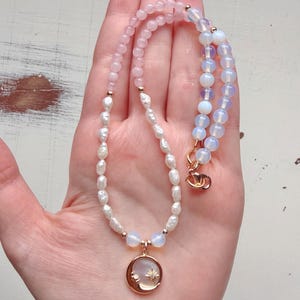 Op de afbeelding: Een kralenketting met een mix van lichtroze ronde kralen, witte onregelmatige zoetwaterparels en doorschijnende opaalachtige kralen. De ketting heeft een goudkleurige ronde hanger met een halve maan en een klein sterontwerp.