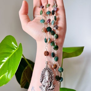 Puede incluir: Un collar de plata con un colgante de Ganesha detallado y cuentas de ágata verde, marrón y púrpura.