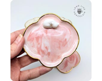 May include: A set of three pink and white marbled ceramic trinket dishes with gold rims. Each dish is shaped like a flower with five petals. The top dish has a small white ceramic cat figurine on top.