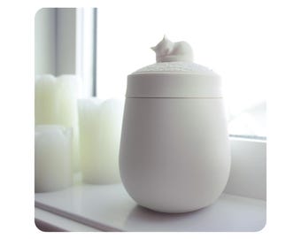 Puede incluir: Un tarro de cerámica blanco con tapa, adornado con una figura de gato. El tarro tiene forma redondeada y está colocado sobre una superficie blanca. La luz suave de una ventana ilumina la escena, con velas blancas al fondo.