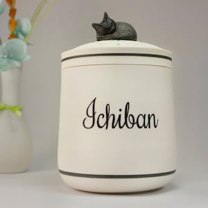 May include: White ceramic jar with a black cat figurine on the lid. The jar has a black stripe around the top and bottom and the word "Ichiban" written in black cursive on the front.