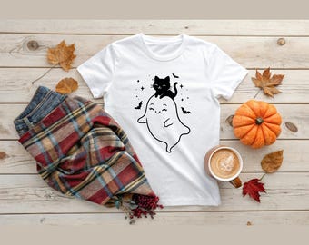 May include: White t-shirt featuring a black graphic of a smiling ghost with a black cat on top. The design includes bats and stars. The shirt is displayed with fall-themed decor, including a pumpkin and leaves, suggesting a Halloween theme.
