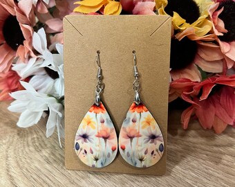 May include: A pair of teardrop-shaped earrings with a floral print. The earrings are made of wood and have silver hooks.
