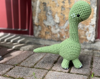 May include: A green crocheted dinosaur toy with black eyes and a long neck. The dinosaur is standing on a brick patio.