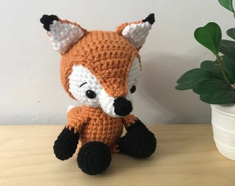 May include: A crocheted orange and white fox plush toy with black eyes and nose, sitting on a wooden surface next to a potted green plant.