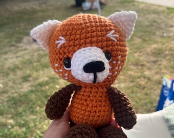 May include: A handmade crochet red panda plush toy. The toy has an orange body, white face and ear accents, and dark brown arms and legs. The plush is held in a person's hand.