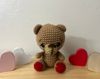 May include: A brown crocheted teddy bear with red crocheted feet sits on a wooden surface with three wooden hearts. One heart is pink, one is white, and one is red.