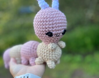 May include: A pink and beige crocheted amigurumi toy shaped like a caterpillar with blue ears. The toy is being held in a person's hand.