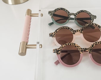 May include: Three pairs of round sunglasses with leopard print frames. The sunglasses are stacked in a clear acrylic tray with a gold handle.