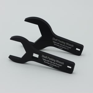 May include: Two black shaft packing wrenches are displayed against a white background. Each wrench has a curved head and a rectangular opening in the handle. Text on the handle reads "Shaft Packing Wrench".