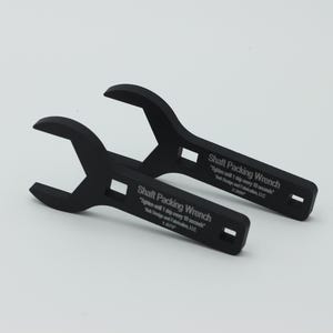 May include: Two black shaft packing wrenches are displayed against a white background. Each wrench has a curved head and a rectangular opening in the handle. Text on the handle reads "Shaft Packing Wrench" and instructions.