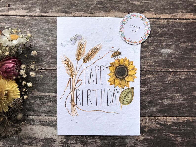 May include: A white card with a sunflower, wheat stalks, a bee, and butterflies. The card says "Happy Birthday" and has a small circle sticker that says "Plant Me".