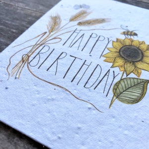 May include: A white card with a sunflower, wheat stalks, and a bee. The card says "Happy Birthday" in black script.