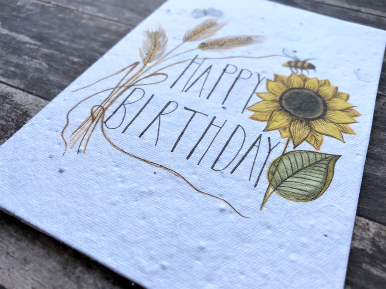 May include: A white card with a sunflower, wheat stalks, and a bee. The card says "Happy Birthday" in black script.